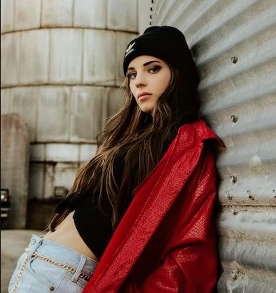 A woman leaning on a wall in a red jacket, black shirt and blue pants with a chain belt